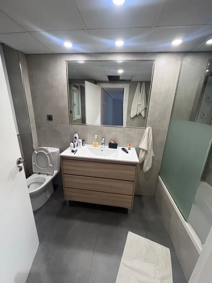 Foto real de un baño antes de la remodelación: lavatorio con mueble de madera, inodoro, tina con mampara y mayólica gris